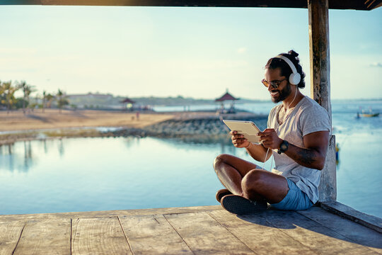 Relaxed and cheerful. Work and vacation. Outdoor portrait of happy young african man using tablet computer on deck near the sea. - Powered by Adobe