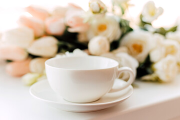 Fototapeta na wymiar A white cup and saucer stand near the flowers on a light background