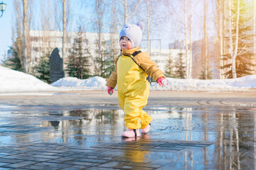 A cute kid 2-3 years old dressed in a yellow waterproof jumpsuit runs through a puddle in a spring...