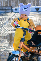 Toddler aged 2-3 years in a yellow waterproof jumpsuit plays with a bell located on the handlebars of a tricycle standing on the cobblestones in a spring park against the background of melting snow
