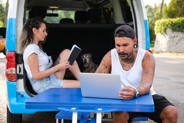 Young tattooed couple reading a book and working on a laptop, sitting in a picnic table and in the...