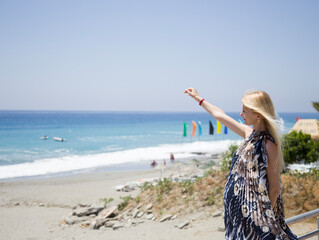 A blonde girl in a short plisse dress strolls along the promenade of the Mediterranean Sea in sunny...
