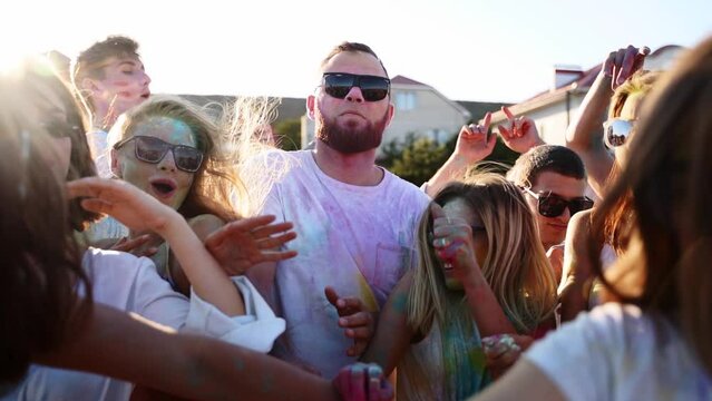 Handsome man with strong face threading way through crowd, starts shouting. Guy gets two handfuls of colored powder in mouth by beautiful girls to have fun at Holi festival. End of covid isolation