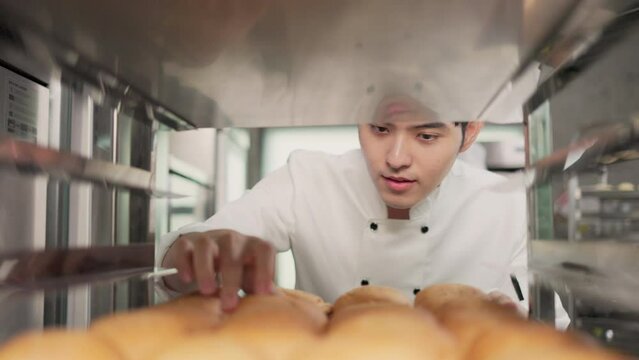 Smiling bakers looking at camera.Chef  baker in a chef dress and hat, cooking together in kitchen.Young asian man takes fresh baked cookies out of modern electric oven in kitchen.
