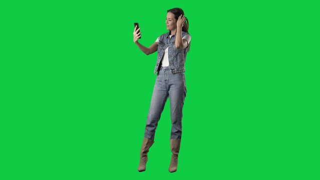 Stylish girl in jeans and boots taking selfies with phone posing and touching hair. Full body isolated on green screen background