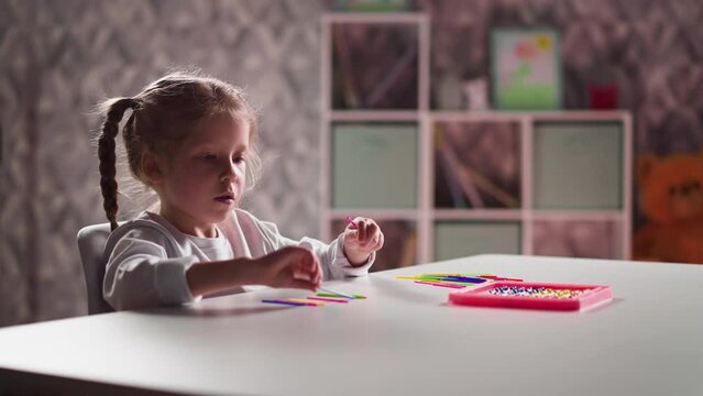 Concentrated little girl student with plaits puts small colorful sticks learning to count at mental arithmetic class in room closeup slow motion
