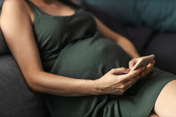 A woman with a big belly is resting and using the phone while sitting on a sofa in the living room