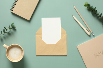 Envelope with blank card inside, cup of coffee, paper notepad, weekly planner, eucalyptus leaves on green table. Flat lay, top view, copy space.