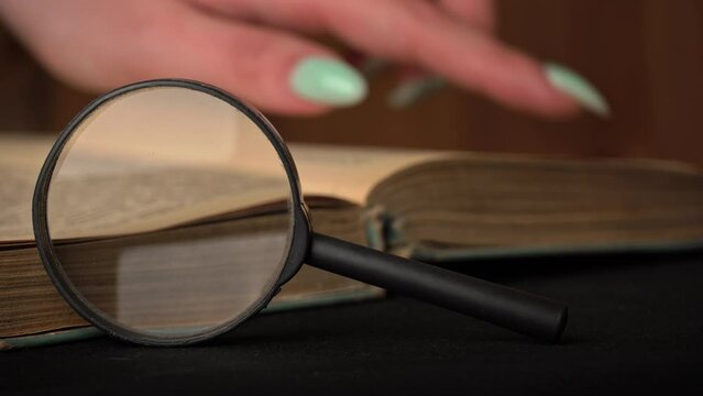 Girl turns the page of an old book next to a magnifying glass. A vintage book with dusty pages lies on a wooden background. Old book and lens. Reading an old library.