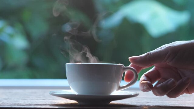 Hands holding a cup of hot coffee or tea to drink in the morning sunrise. Soft steam smoke floated above the white coffee cup on the wooden table, put on a saucer. Hot coffee drinking concept.
