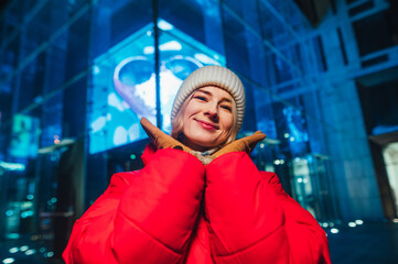 Cute lady in a hat and red jacket stands against the backdrop of a blue light building and looks at the camera with a smile on his face.