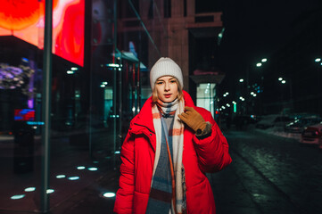 Night portrait of a young woman in warm clothes on the streets of the metropolis while walking, looking at the camera.