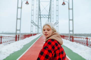 Beautiful woman with blond hair walks on a snowy bridge with a serious face looking at the camera and posing against the backdrop of winter urban views.