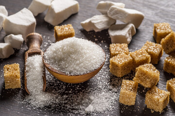 Granulated table sugar in wooden bowl and in the spoon and sugar cubes around it on the table.