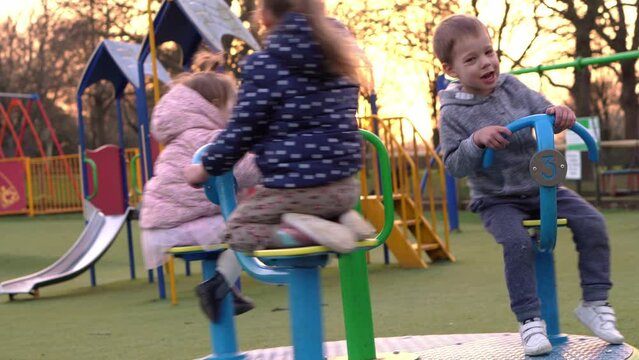 Three little Happy Children having fun spinning on merry-go-round. Smiling Siblings friends playing outside. emotional kids rides on carousel at playground nursery in park.Happy family, childhood