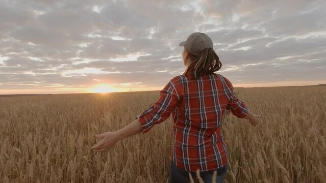 Successful woman farmer walking in wheat field to meet sun at sunset raising upper hands with happiness.
