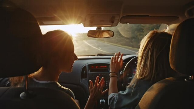 Rear view of cheerful young girlfriends singing their favourite song and dancing while driving at sunset. Two beautiful women travelling and having good time together . Concept of friendship