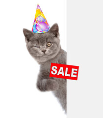 Winking kitten wearing sunglasses and  birthday cap looks from behind empty white banner and holds sales symbol. isolated on white background