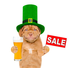 St Patrick's Day concept. Smiling puppy wearing hat of the leprechaun holds glass of beer shows sales symbol. isolated on white background