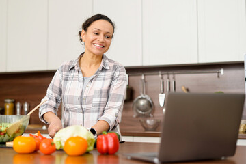 Cooking at home. Mid-age female is watching cooking video recipes on a laptop and cutting salad. Housewife preparing food and watching TV series