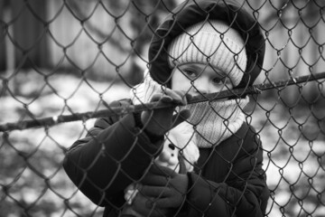 Little refugee girl with a toy behind a metal fence. Social problem of refugees and internally displaced persons. Russia's war against the Ukrainian people - obrazy, fototapety, plakaty