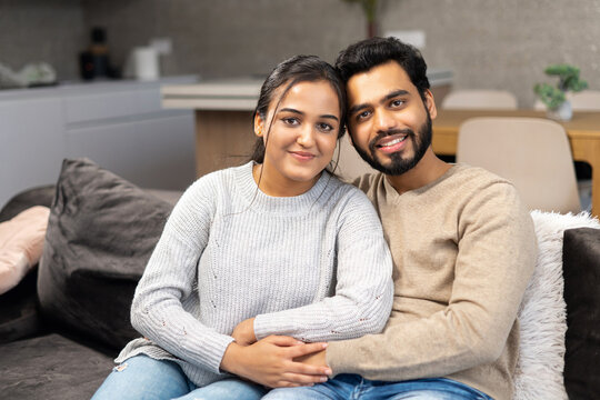 Multi ethnic couple in love rent a house together. Happy Indian newlyweds moved in new apartment, sitting at the sofa in embraces, looking at the camera