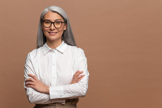 Positive sanguine senior woman smiling cheerfully while posing arms crossed. Hilarious mature asian gray-haired lady standing isolated over beige background