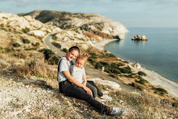 Naklejka na ściany i meble family, little girl sitting in mom's arms on the background of rocky sea shore, sunny day, positive emotions