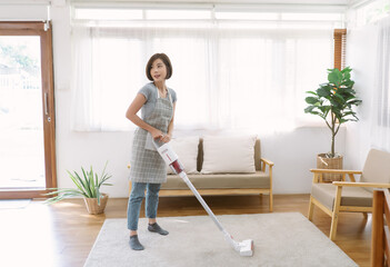 Happy beautiful young Asian woman doing house chores in apron using vacuum cleaning the carpetin a...