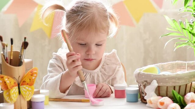 A two-year-old girl with two tails paints an Easter egg with pink paint. Happy Easter.