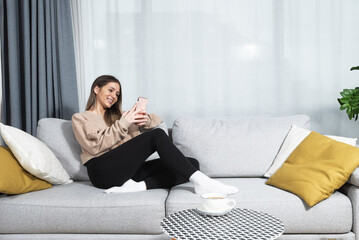 Smiling teenager girl relax lying on sofa at home using cellphone texting chatting with friend,...