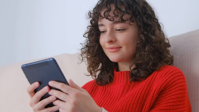 Happy Ukrainian woman reading ebook on tablet computer at home. Cheerful white female with curly hair browsing internet on modern portable gadget