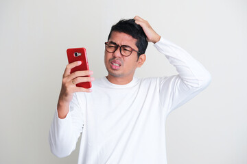 Adult Asian man scratching his head and showing confused expression while looking to his mobile...