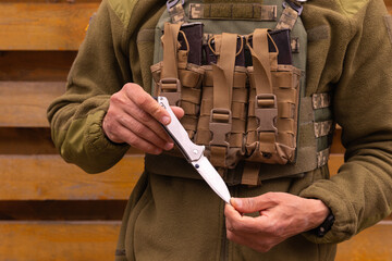 A military man in a bulletproof vest holds a folding tourist tactical knife in his hands.