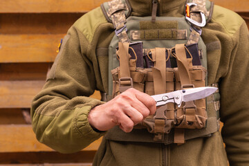 A military man in a bulletproof vest holds a folding tourist tactical knife in his hands. - obrazy, fototapety, plakaty