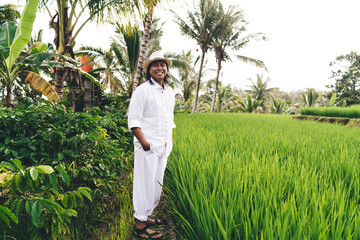 Cheerful Balinese entrepreneur in hat and white wear smiling while visiting own farmland with rice fields and coffee bushes - enjoying business lifestyle at countryside of Indonesia, happiness