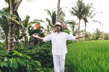 Cheerful adult farmer in white clothes and hat smiling near rice fields and coffee bushes in...