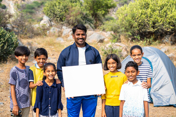 Smiling teacher with teenage kids showing white empty board by looking at camera at mountain top - concept of advertisement, trekking and promotion for summer camp.