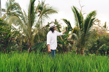 Happy male tourist in hat clicking selfie images via modern cellular gadget using 4g wireless while walking at rice fields at Ubud, cheerful man enjoying online calling for communicate at Bali
