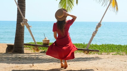 Back view of woman in red dress and hat sit on swing at tropical beach with fresh opened coconut, palm tree branch, turquoise sea on background. Sunny hot summer day, rest, holidays, vacations, travel