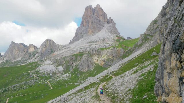 A young couple walks a high mountain trail with sports but non-technical equipment, they are surrounded by bare rock peaks. a shelter stands out on a peak in the background