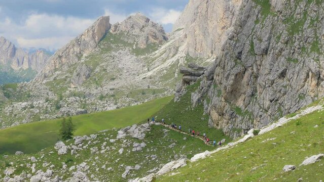 A group of people walk in a row on a high mountain path that winds through the green grass. They all wear sports gear. All around bare rock mountains stand out on a sunny day