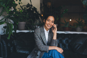 Portrait of happy Asian woman in jacket smiling at camera while calling to friend for talking indoors, successful female enjoying mobile communication using smartphone application and wireless