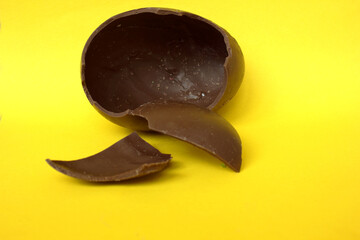 A broken chocolate egg on yellow background, close-up. A chocolate Easter egg with a broken top. Happy Easter. Chocolate. Sweets for children and adults. Candy. Dessert. Copy space. Top view. Flat lay
