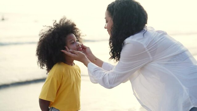 Happy African family mom and child girl kid enjoy and fun outdoor lifestyle on beach vacation. Mother applying sunblock to little daughter while walking together on tropical beach at summer sunset.