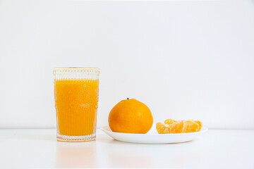 Glass of natural orange juice besides a full orange and peeled orange pieces on a dish, on a white table, against white background