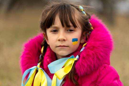 A little Ukrainian refugee girl with a Ukrainian flag painted on her cheek is sad about her lost home. Stop the war in Ukraine. Help save the Ukrainian nation