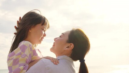 Happy Asian family on beach vacation. Mother carrying and kissing little daughter while walking on tropical beach at summer sunset. Mom and child girl kid enjoy and fun outdoor lifestyle on the beach
