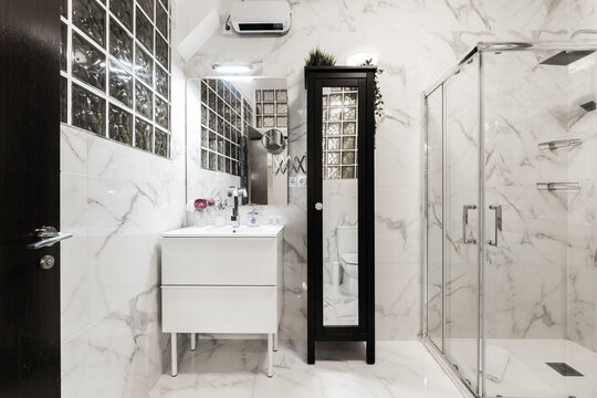 Bathroom with shower cabin with glass partition, frameless rectangular mirror, long black wooden cabinet with mirror and gray veined marble clad walls in combination with cobblestone wall