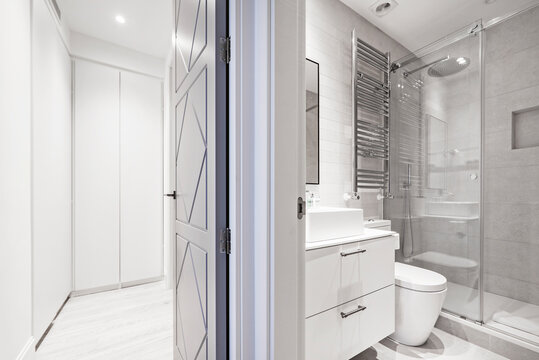 Bathroom with white resin sink, marble walls, white wooden furniture, shower cabin with screen, chrome towel dryer radiator and access to a dressing room